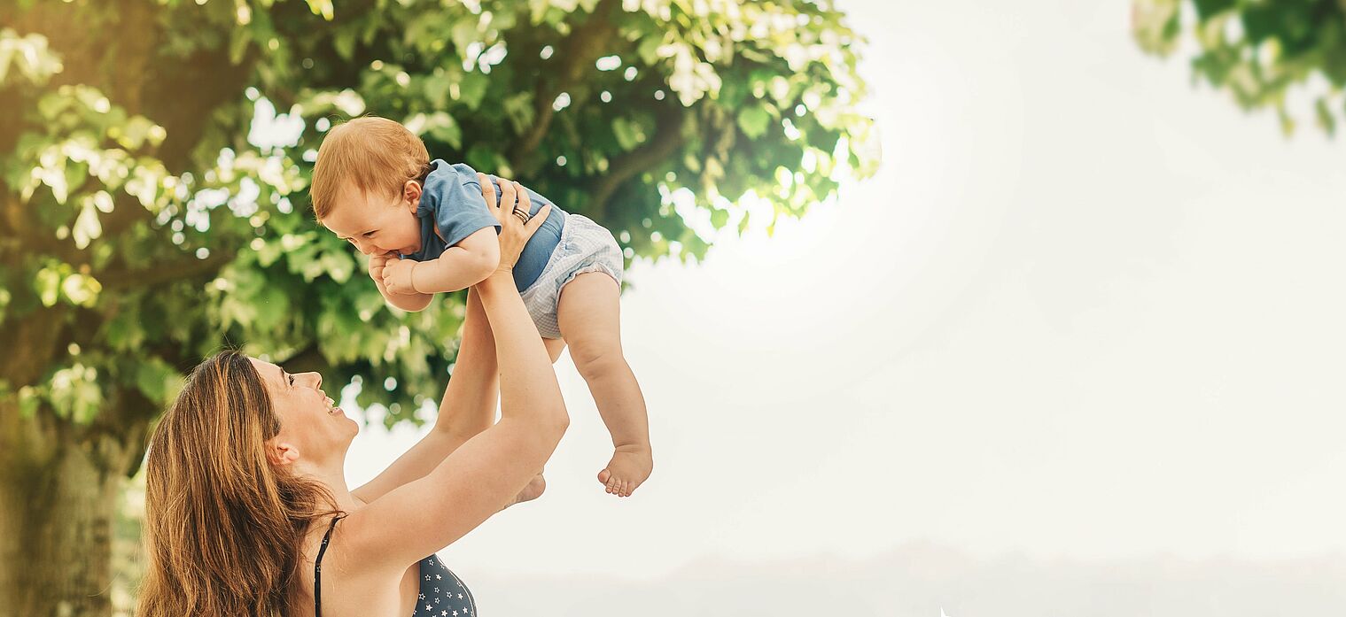 Mother lifting her baby joyfully in the air outdoors, with green trees in the background and both smiling.